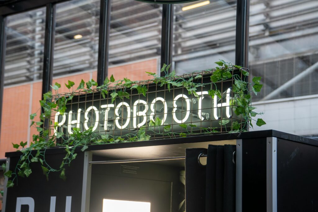 A contemporary photobooth with neon sign and greenery, set outdoors.